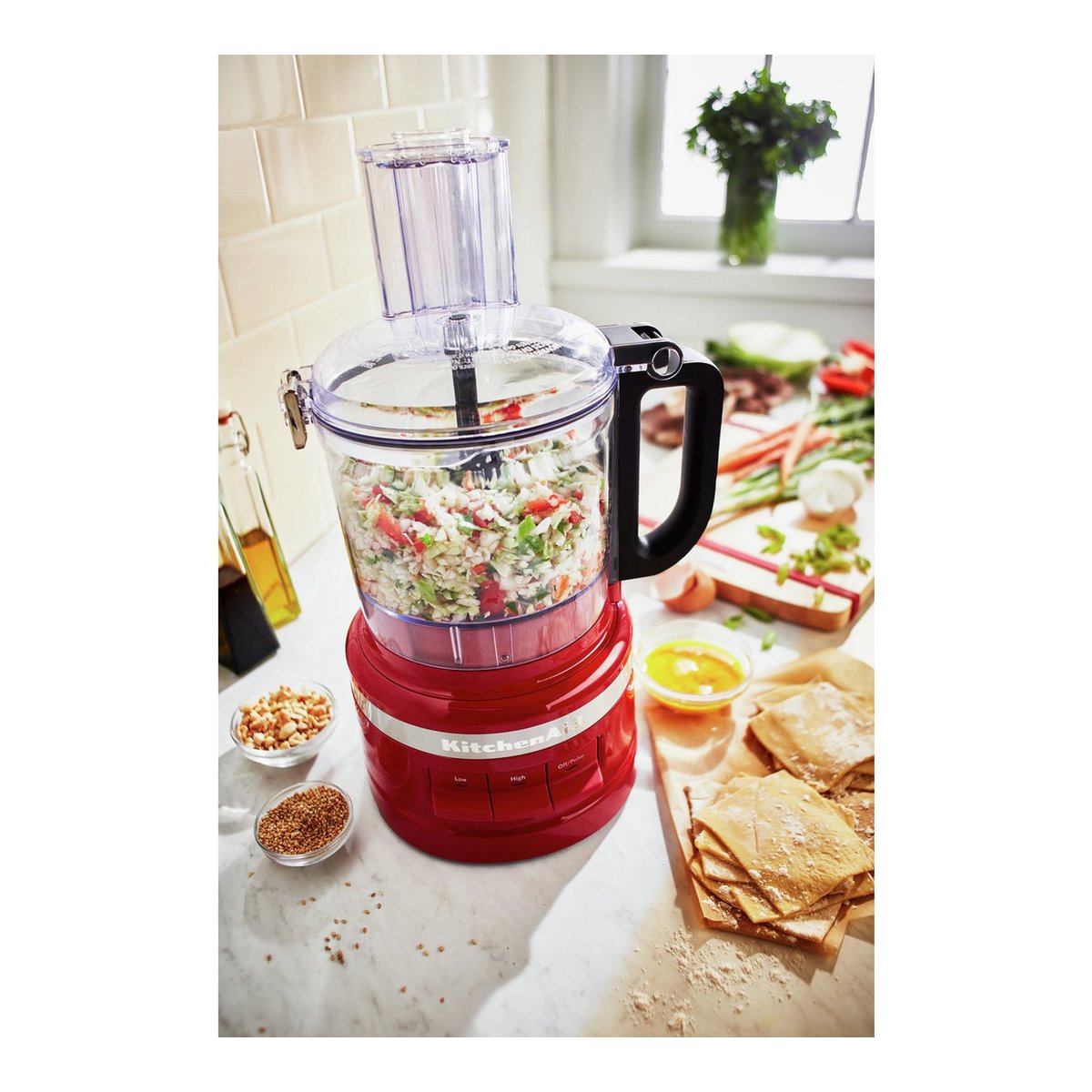 Compact KitchenAid food processor in Empire Red with a clear bowl, black handle, and various chopped vegetables inside, surrounded by ingredients on a marble countertop.