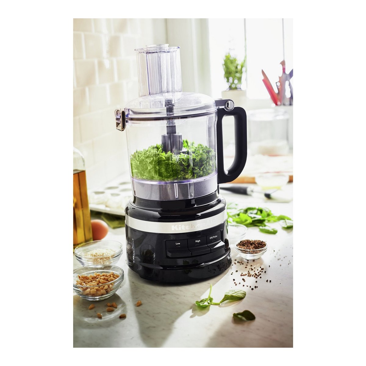 Compact KitchenAid food processor in matte black with a transparent bowl, filled with chopped greens, featuring a sleek design and control buttons on the base, set on a marble countertop.