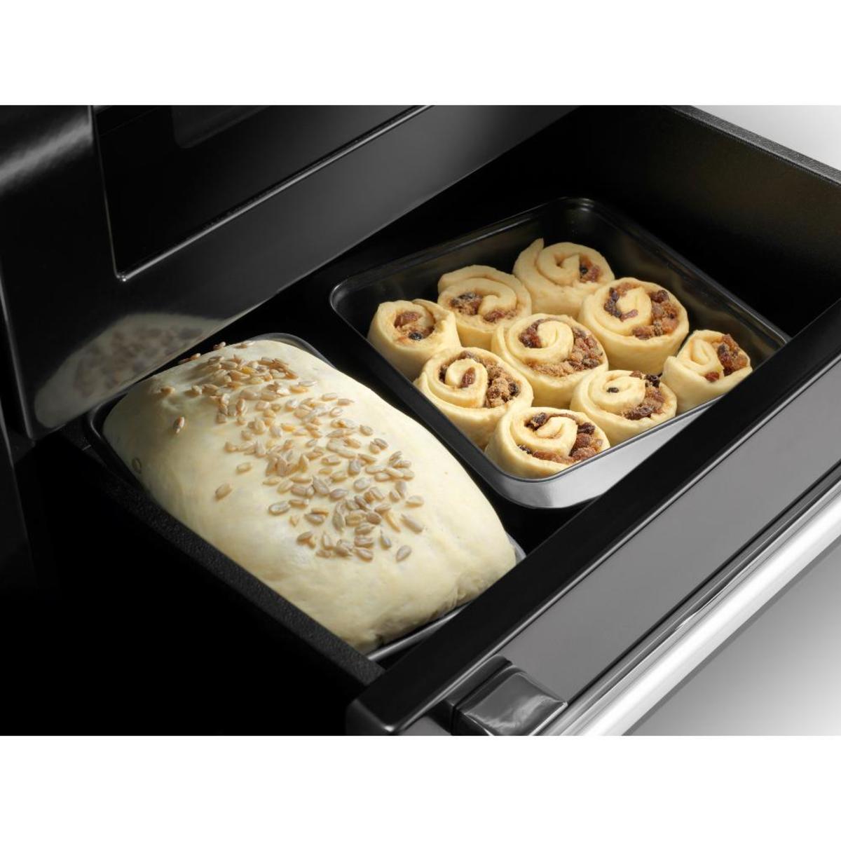 Deluxe 110cm induction range cooker in slate with chrome trim, featuring a baking tray with cinnamon rolls and a loaf of bread in a black oven compartment.