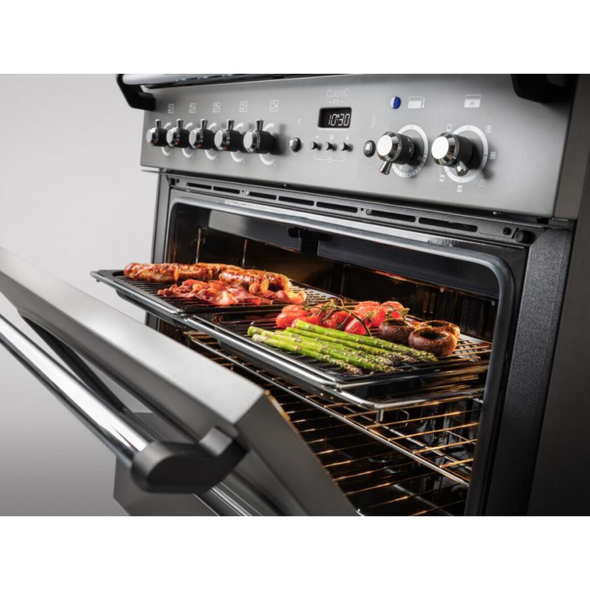 Classic FX 90cm induction range cooker in slate with brass trim, featuring multiple knobs, a large oven cavity, and a tray of grilled meats and vegetables inside.