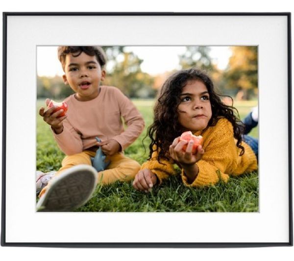 Two children sit on green grass, one wearing a light brown long-sleeve shirt and the other in a yellow sweater, both holding fruit and looking thoughtfully at the camera.