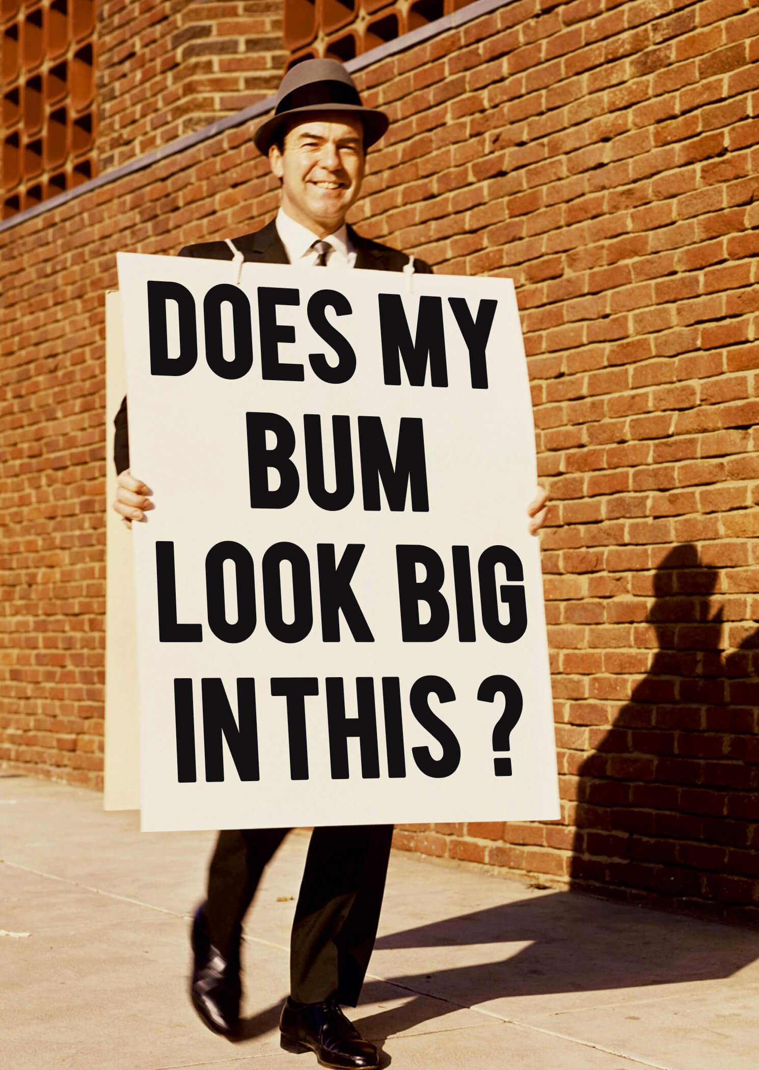 A man in a black hat and suit holds a large sign with bold black text reading "DOES MY BUM LOOK BIG IN THIS?" against a brick wall background.