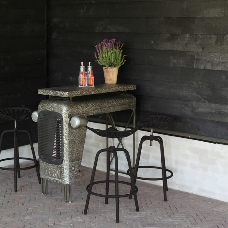 Black Esschert Design Bar Tractor Chair features a textured metal seat resembling a tractor, paired with sturdy black metal legs, set against a rustic black wall and potted plant.