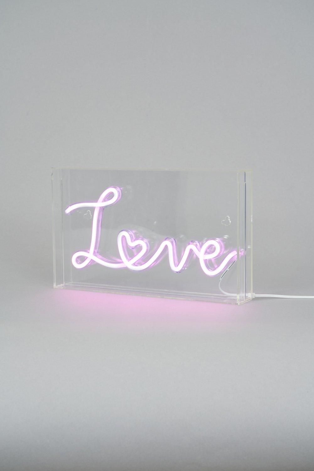 Pink neon light box with the word "Love" in cursive, set in a clear acrylic frame, emitting a soft pink glow from the base.