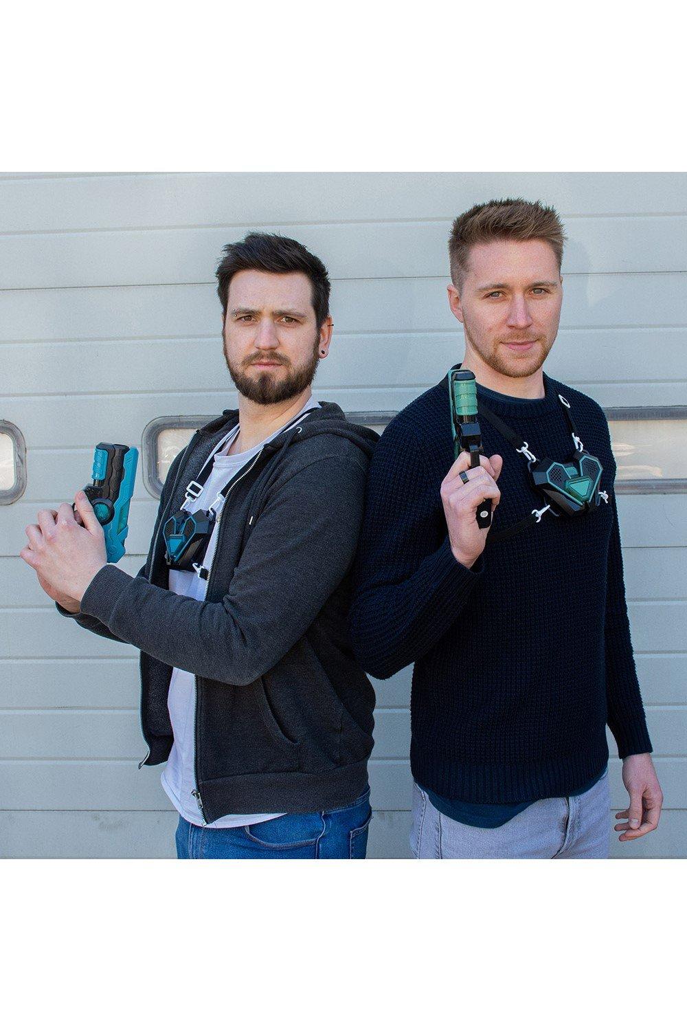 Two men stand back-to-back, each holding a laser tag gun; one wears a grey hoodie and the other a navy sweater, both equipped with electronic vests featuring green accents.