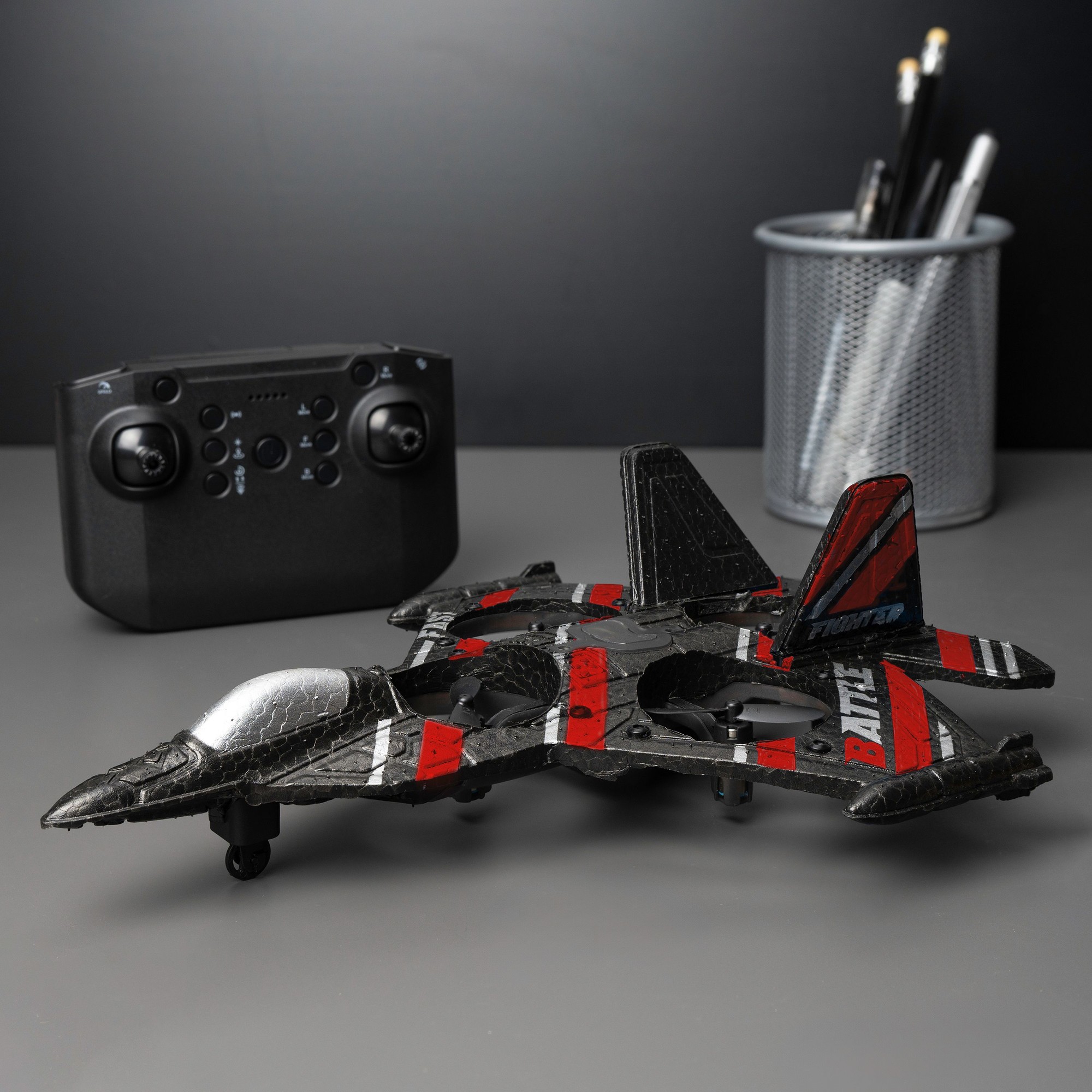 RED5 Jet Fighter Drone in black with red accents, featuring a sleek design, a shiny silver cockpit, and four wheels, alongside a black remote control and a pen holder in the background.