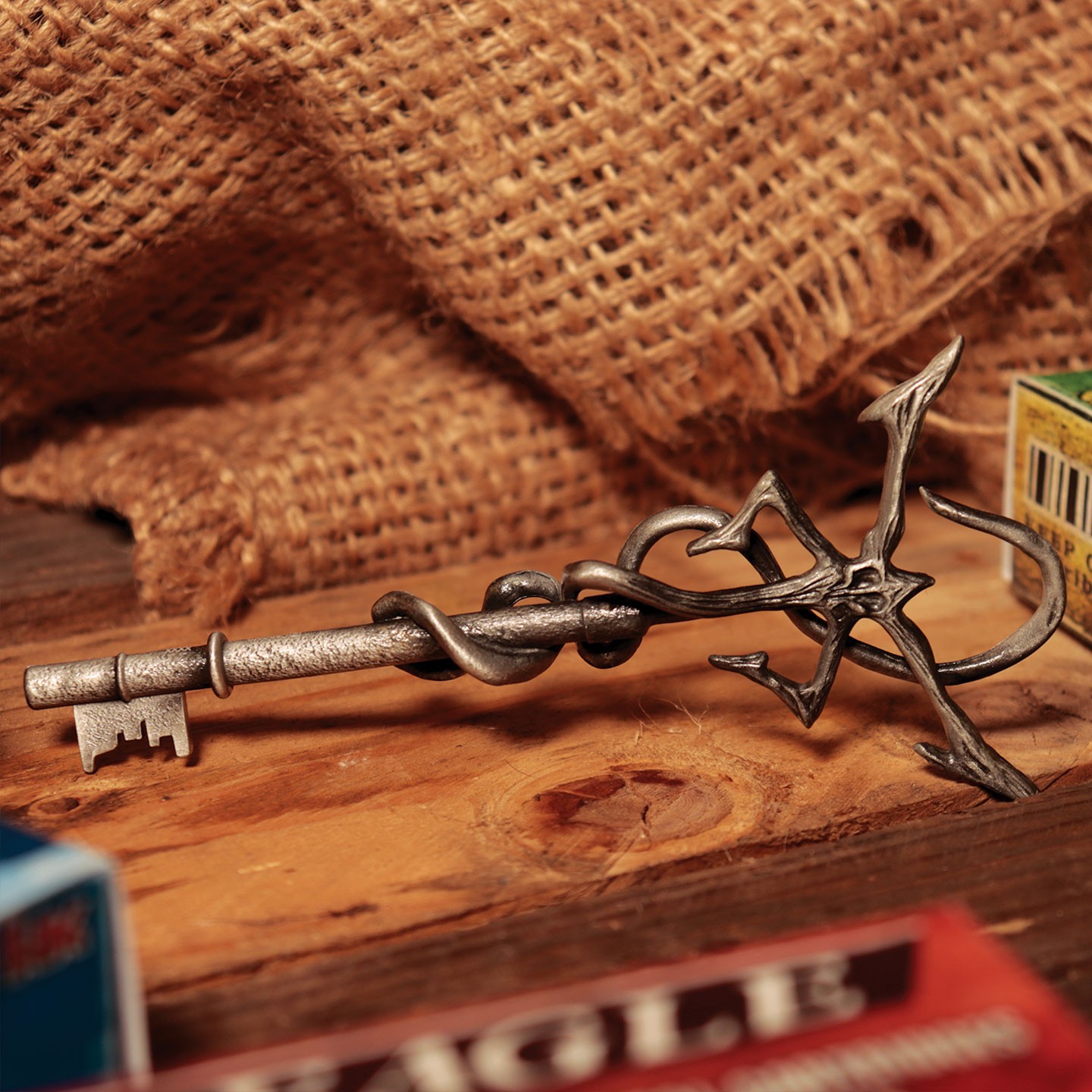A metallic key with an ornate design featuring a twisted vine and insignia, set against a rustic wooden surface and burlap backdrop.