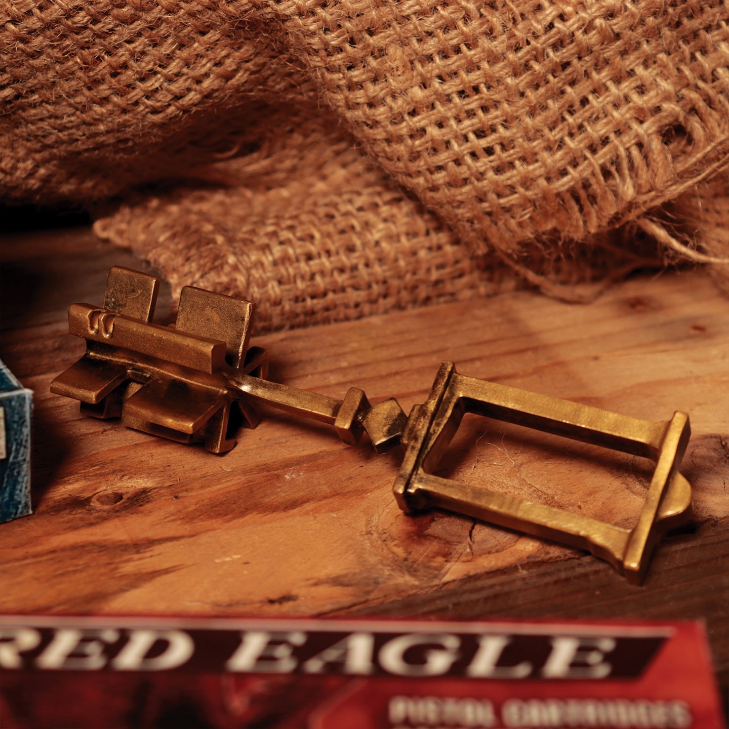 Brass Wayshrine Key from Resident Evil 4, featuring intricate detailing and a vintage design, resting on a wooden surface with burlap and game packaging nearby.