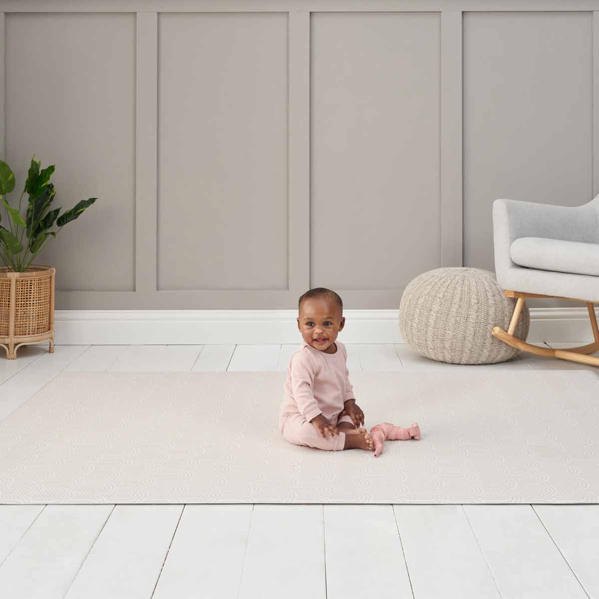 Luxury reversible playmat in soft beige with a textured pattern, featuring a baby sitting on it, surrounded by a neutral room with a plant and cozy furnishings.