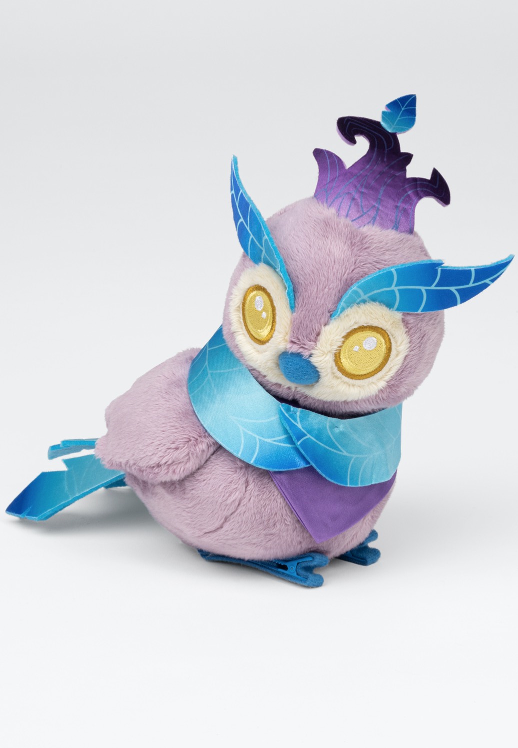 A plush owl toy in soft lavender fur, featuring large yellow eyes, blue feather accents, and a purple crown, adorned with a blue and purple scarf.
