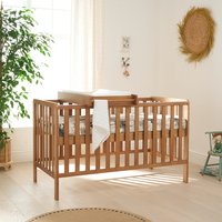 Honey oak cot bed with slatted sides, integrated changer, and mattress in a bright nursery setting.