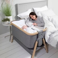 Gray Tutti Bambini CoZee Oak Bedside Crib with wooden legs, mesh sides, and adjustable height next to a bed.