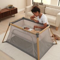 Gray travel cot with wooden frame, mesh sides, and a day & night shade, placed in a living room setting.
