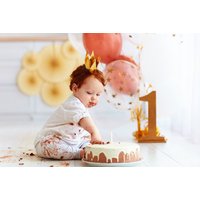 Toddler in white outfit with cake, gold crown, surrounded by balloons and large gold number one in a studio setting.