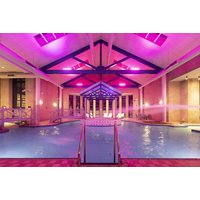 Indoor hotel pool with pink-lit ceiling, wooden beams, and large windows, surrounded by columns and lounge chairs.