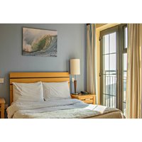 Hotel room with wooden bed, white linens, ocean wave painting, and balcony doors with sea view.