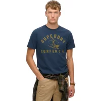 Blue Superdry Lo-fi Outdoor Relaxed T-shirt with yellow surfer graphic and text, worn by a model with beige pants.
