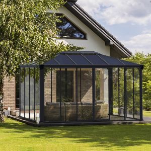 A 10' x 15' Palram Canopia Ledro 4300 enclosed gazebo featuring a black aluminum frame, clear polycarbonate panels, and a modern design, set on a green lawn beside a house.
