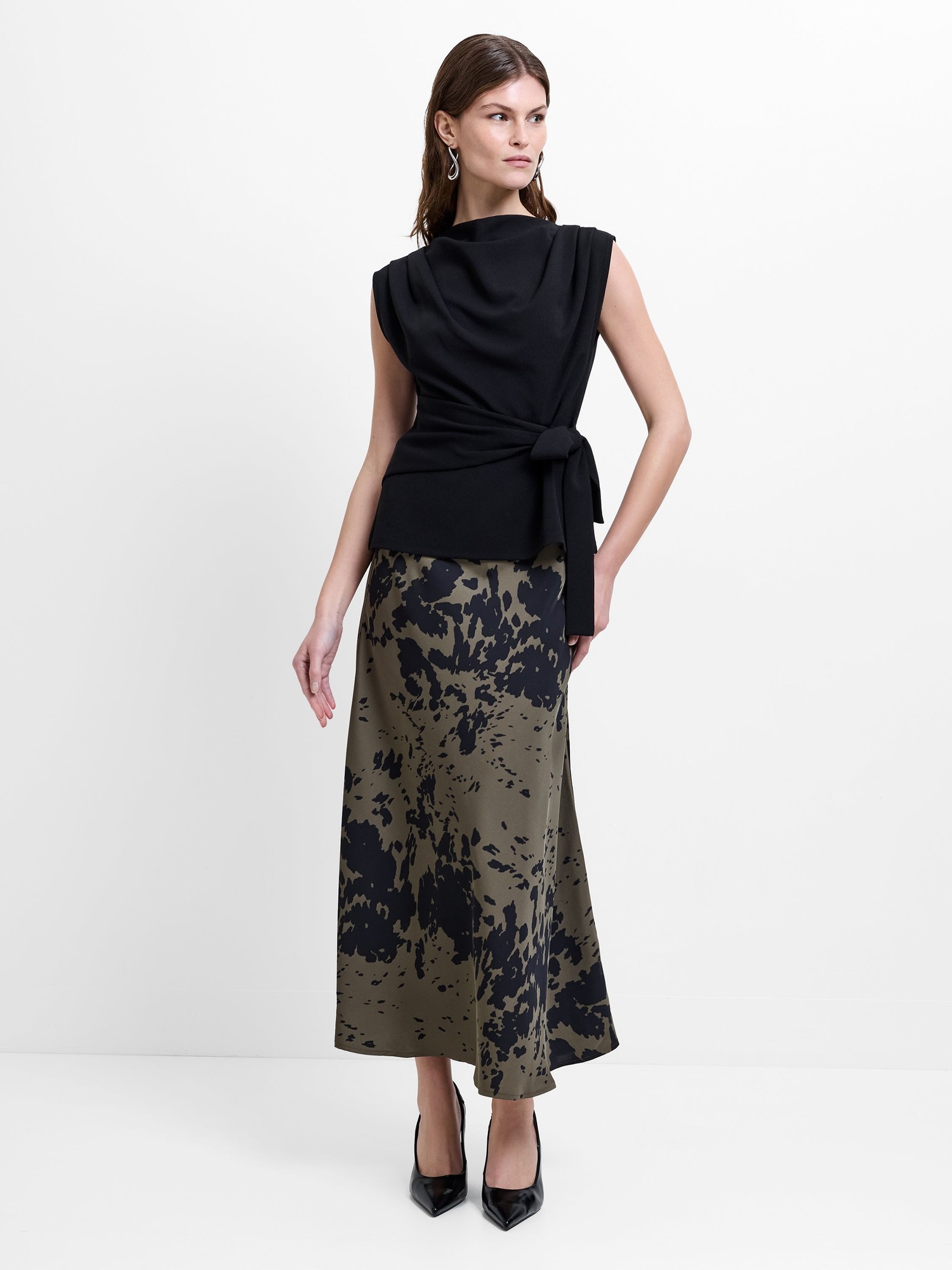 Black satin midi skirt with a flowing silhouette, featuring a unique abstract print in olive and beige tones, paired with a stylish black top and high heels.