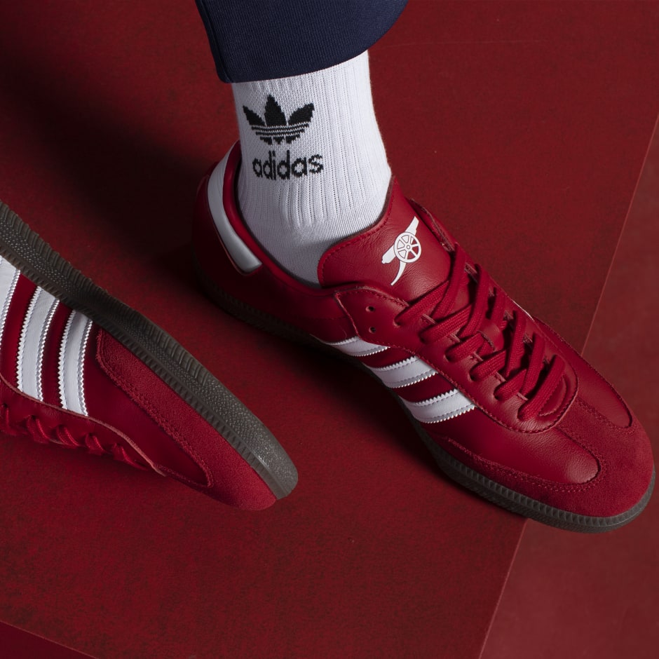 Red leather Adidas Samba shoes featuring white stripes and Arsenal FC logo, paired with white Adidas socks, set against a red background. Size US 10.5.