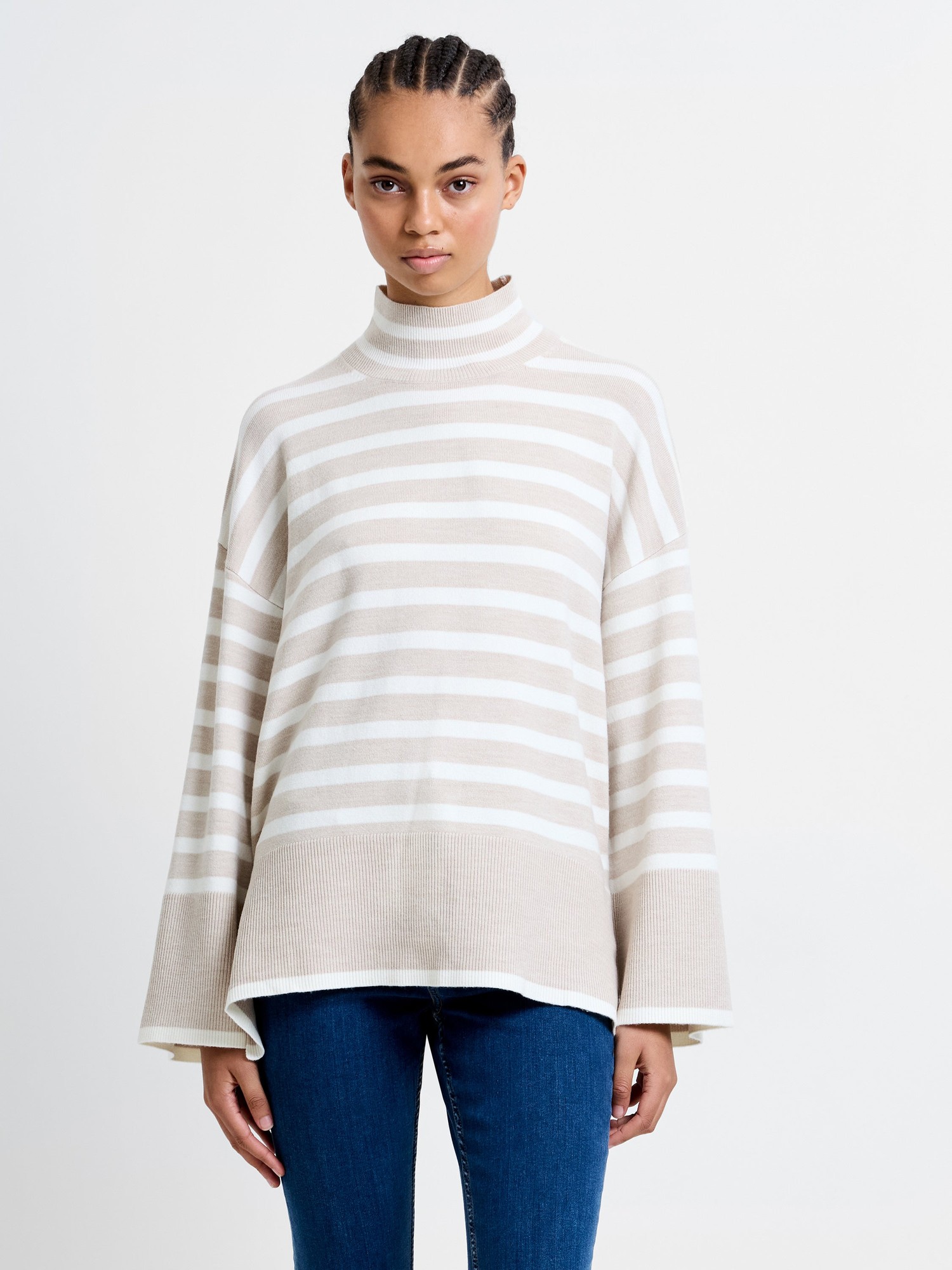 Beige and white striped high neck jumper with side splits, featuring a relaxed fit and ribbed hem, worn with blue jeans.
