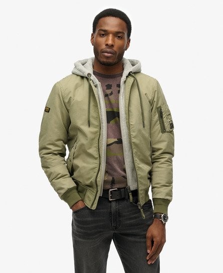Olive green bomber jacket with a gray hoodie lining, featuring a zip front, ribbed cuffs, and a patch on the sleeve, paired with a camo print shirt and dark jeans.