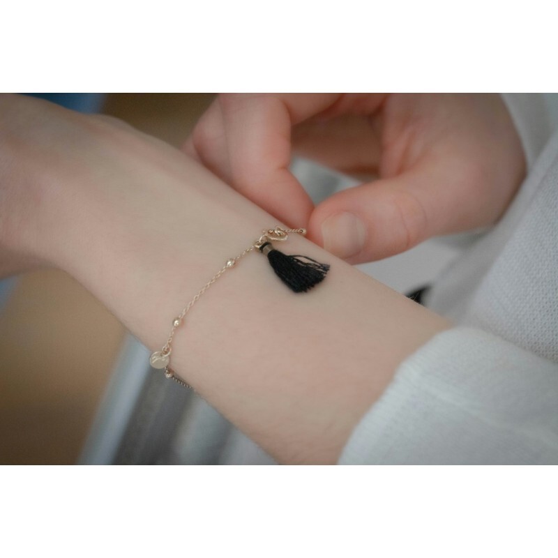 Gold chain bracelet featuring a black tassel charm, worn on a wrist, with delicate beads and a lobster clasp for secure fastening.