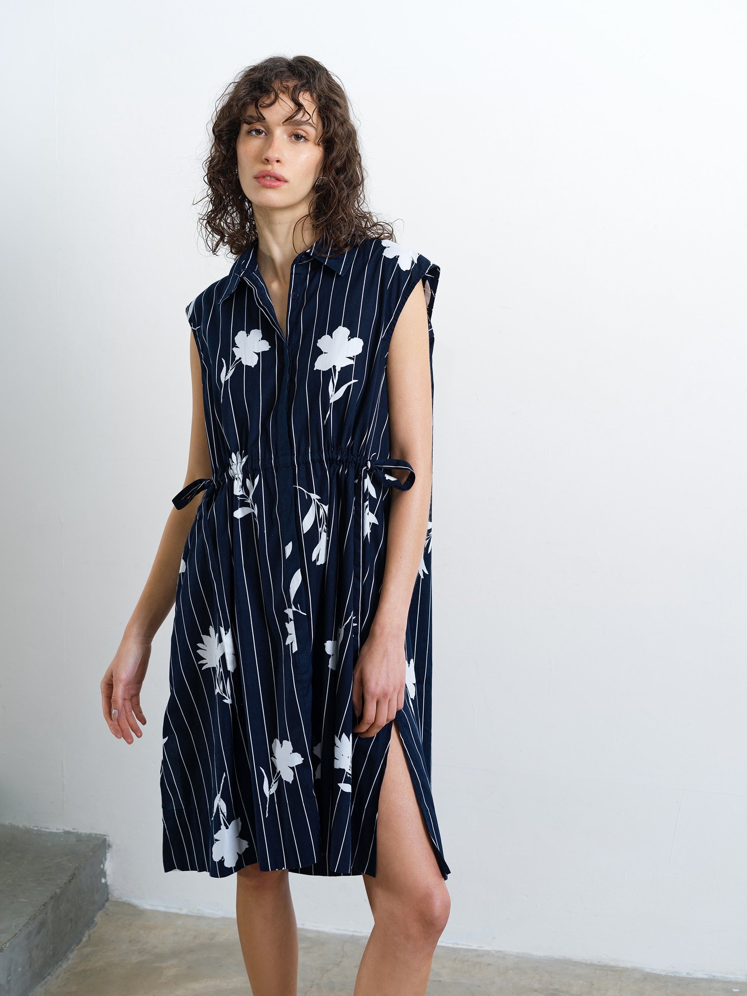 Navy cotton sleeveless drawstring dress with vertical white stripes and floral print, featuring a collar, side slits, and tie details at the waist.