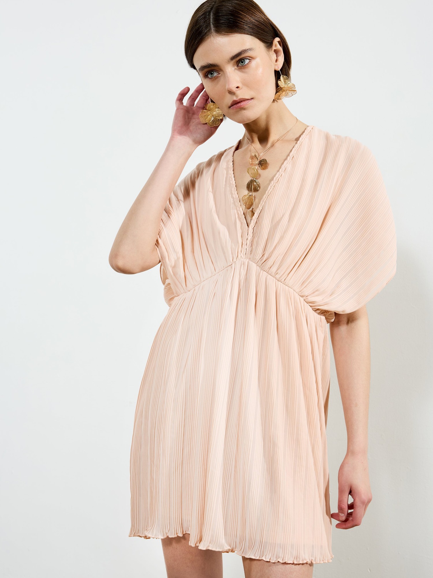 Ballet pink mini dress featuring a V-neck, pleated fabric, and short, flowing sleeves, styled with statement earrings and a delicate necklace.