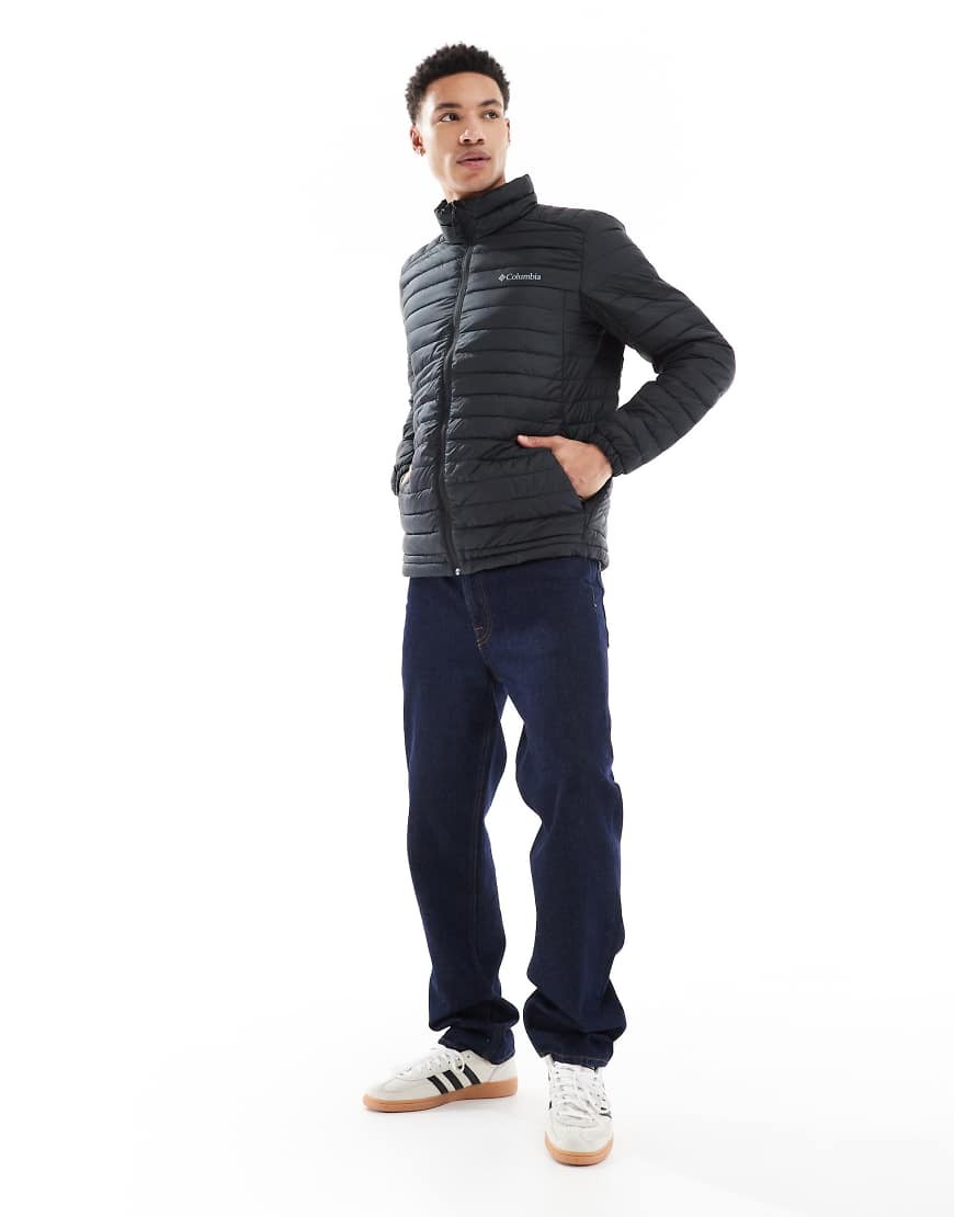 Men's black puffer jacket by Columbia, featuring a zip-up front, high collar, and logo on the chest, paired with blue jeans and white sneakers.