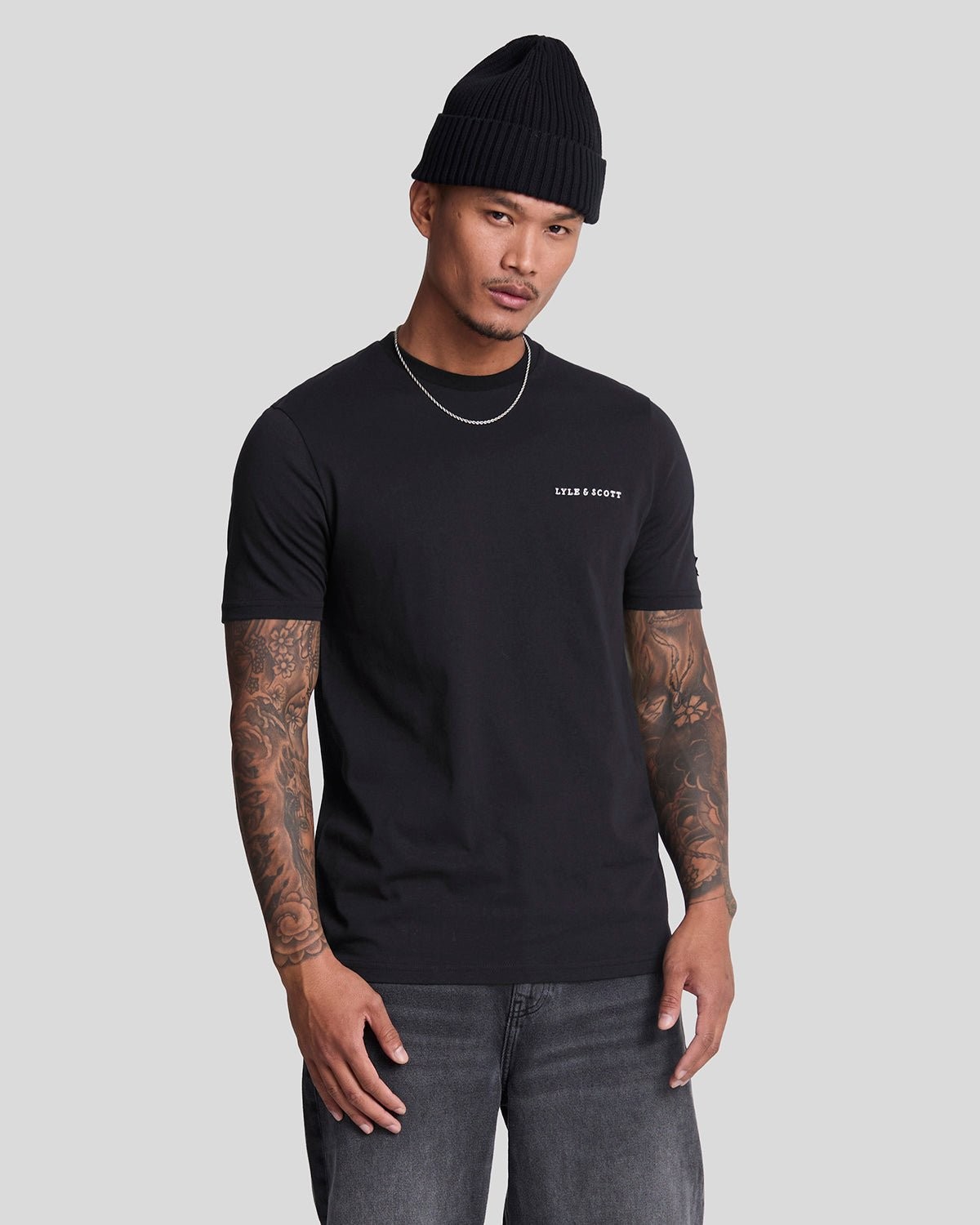 Dark navy cotton t-shirt featuring a white embroidered Lyle & Scott logo on the chest, worn by a model with tattoos and a black beanie.