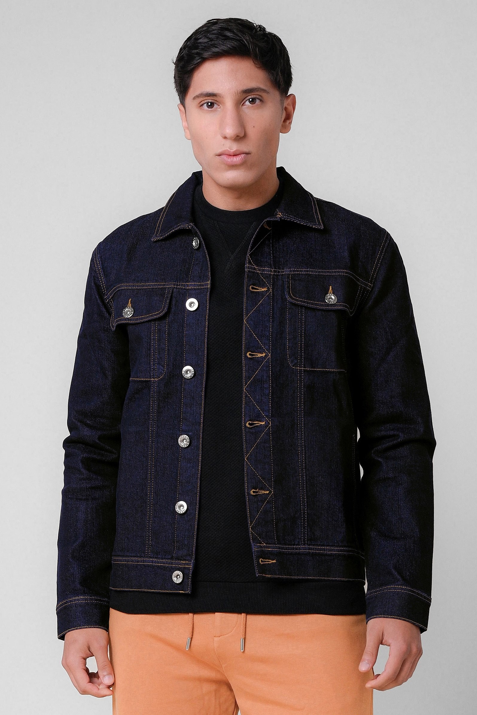 Mens dark wash long sleeve raw denim jacket in deep blue, featuring button closure, classic collar, and chest pockets, styled with a black sweater and light orange pants.