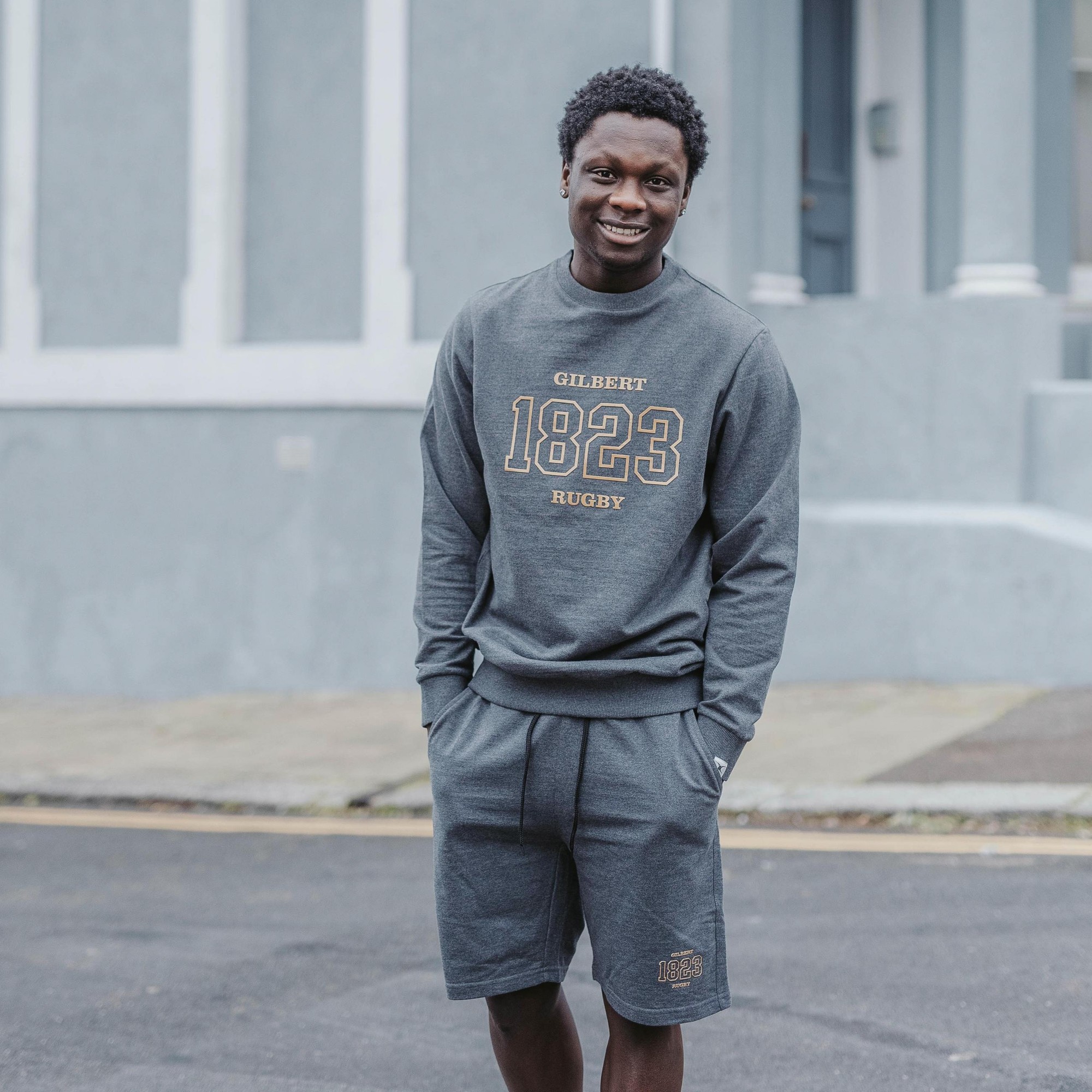 Gray sweatshirt with "GILBERT 1823 RUGBY" printed in gold, featuring a crew neck and ribbed cuffs, paired with matching gray shorts.