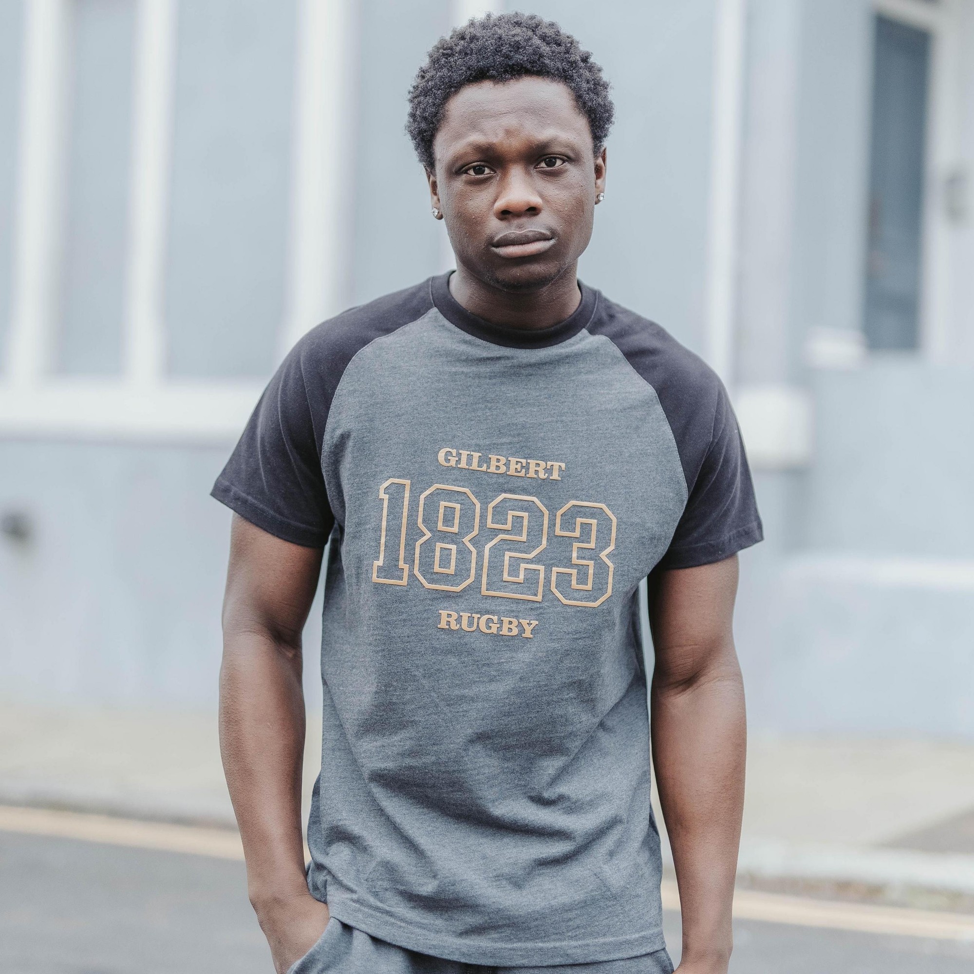 Gray and black short-sleeve leisure t-shirt featuring "GILBERT 1823 RUGBY" in bold gold lettering on the front, made from soft cotton material.