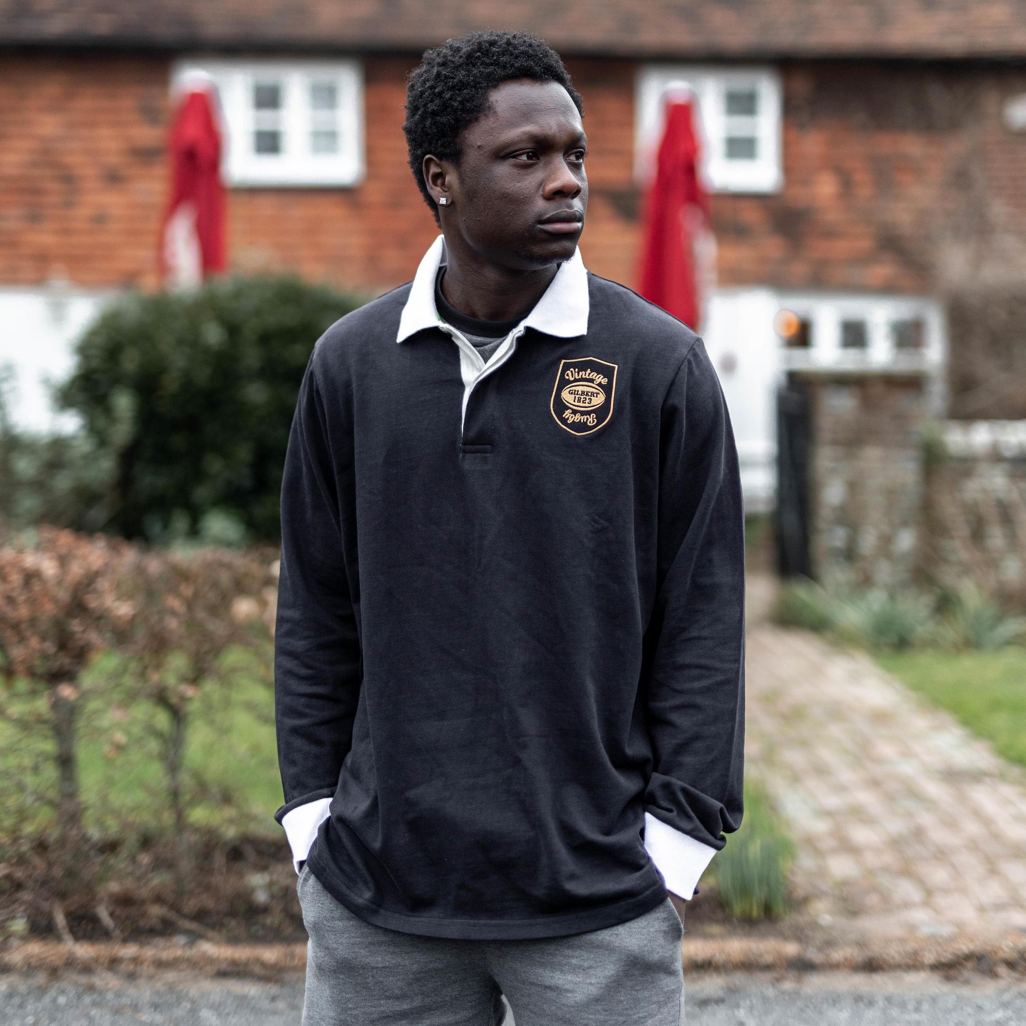 Black vintage rugby shirt with a white collar, featuring a gold embroidered logo on the left chest, made of a soft, durable material, worn by a man outdoors.