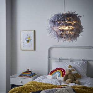 Grey feather light shade with a fluffy, textured appearance, designed to fit easily, illuminating a cozy bedroom setting with a bed, colorful blanket, and decorative items.