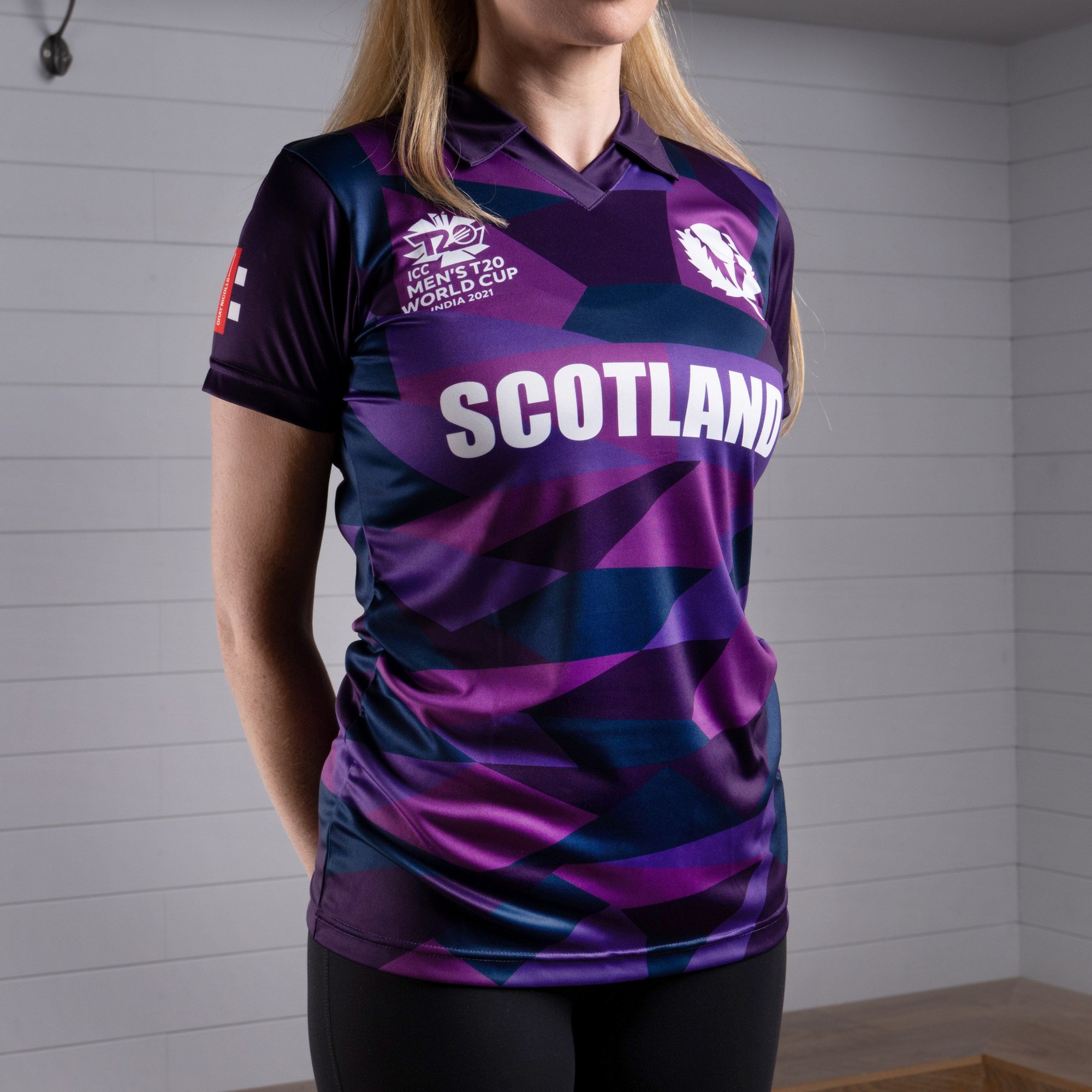 Short sleeve women's cricket shirt in purple and navy, featuring Scotland's logo and "SCOTLAND" text, made from lightweight, breathable material with a modern geometric pattern.