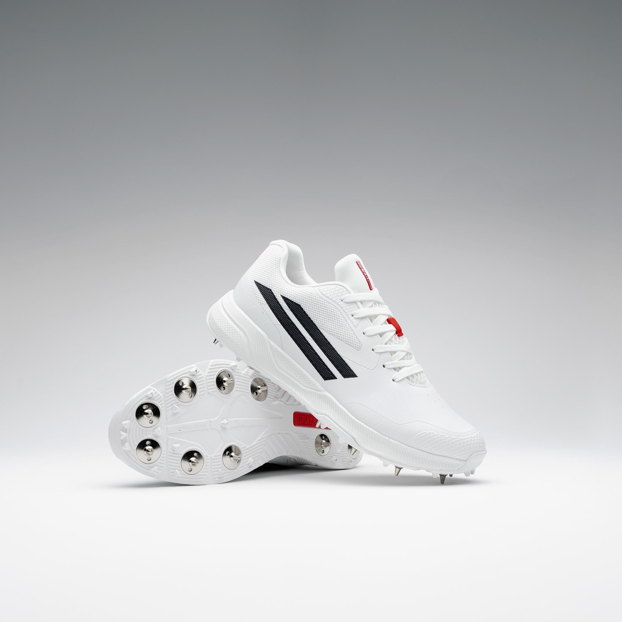 White Gray-Nicolls Revo Pro 3.0 adult spike shoes featuring a textured upper, black stripes, red accents, and a studded outsole for enhanced grip on the field.