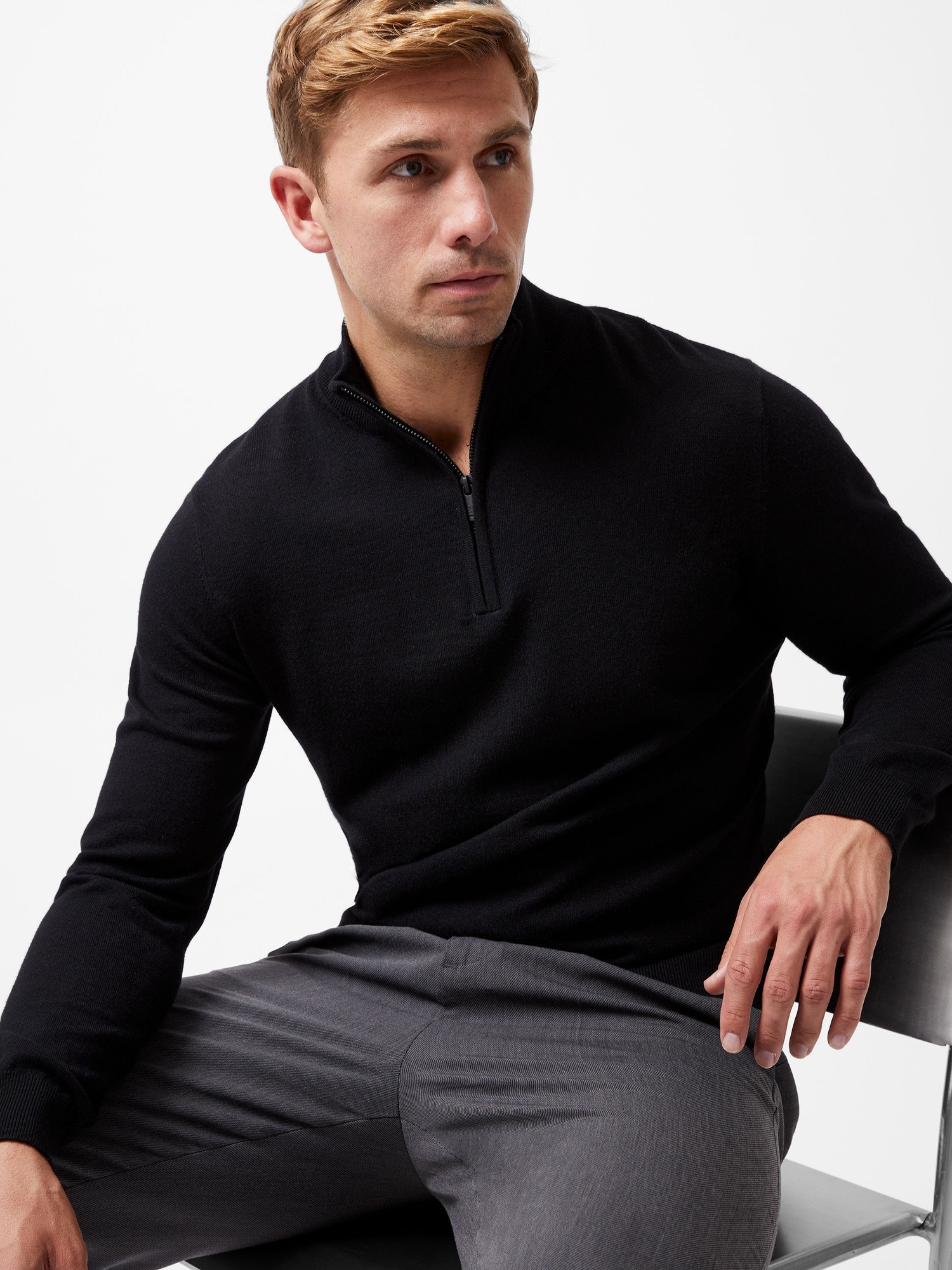 Black half zip knitted jumper made of soft fabric, featuring a stand collar and long sleeves, styled with a relaxed fit, worn by a man seated on a chair.