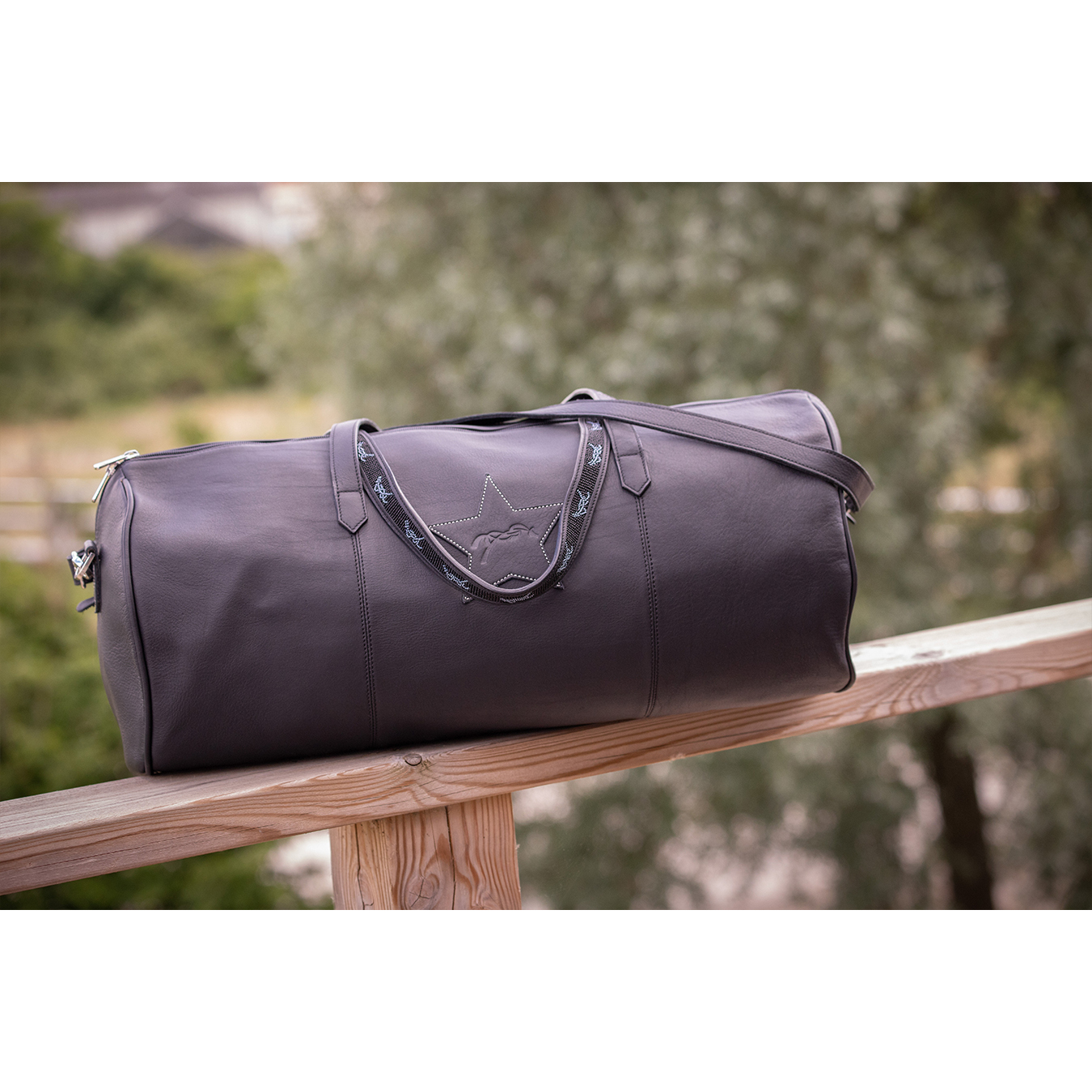 Stylish dark gray leather handbag with a star design, featuring dual handles and a detachable shoulder strap, set against a blurred outdoor background.