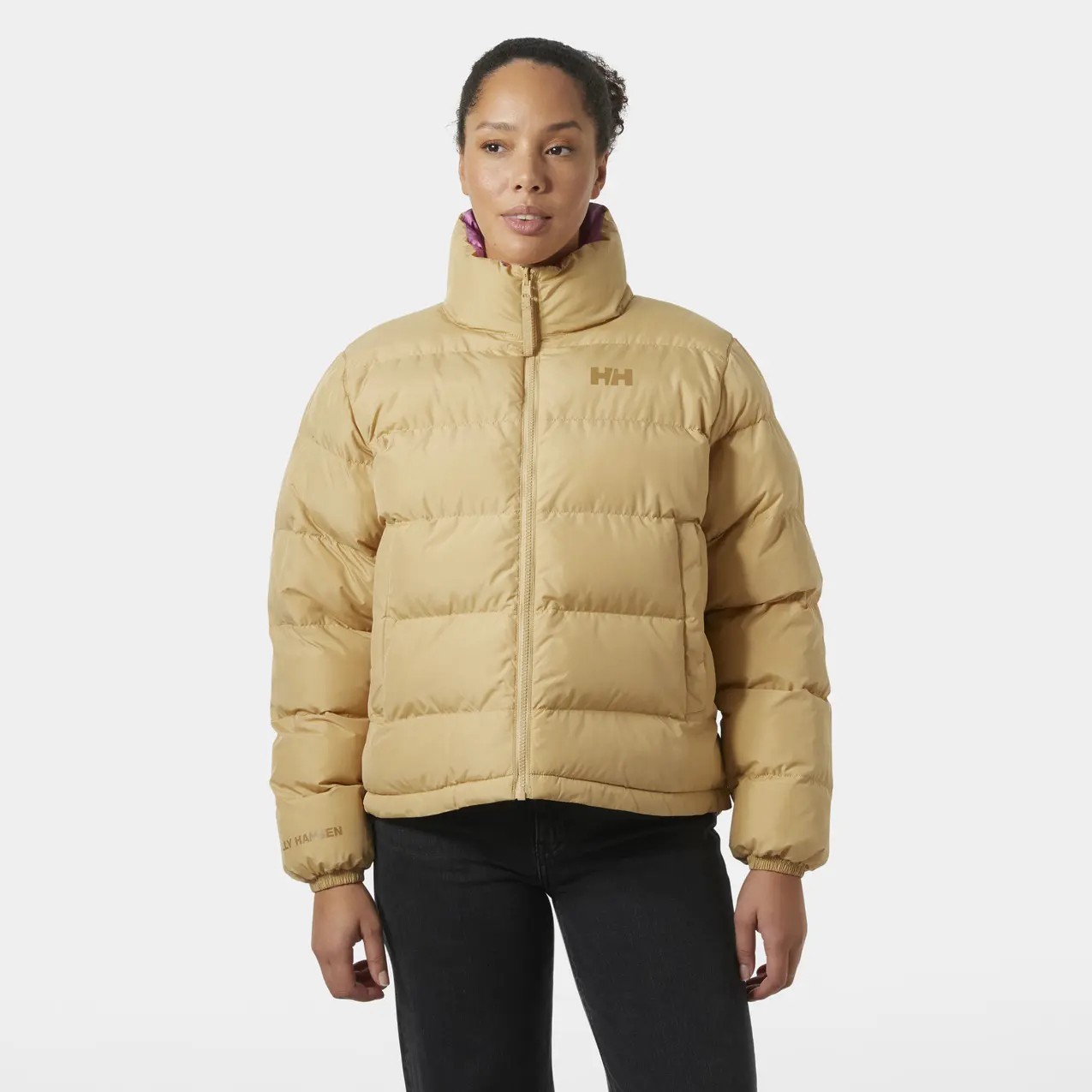 Light tan women's down jacket by Helly Hansen, featuring a high collar, puffy design, and the brand's logo on the chest, paired with dark jeans for a casual look.