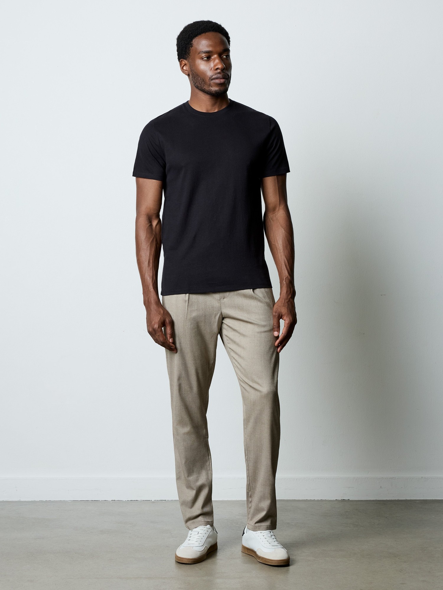Light beige herringbone trousers with a tailored fit, paired with a black t-shirt and white sneakers, showcased on a standing male model against a plain background.