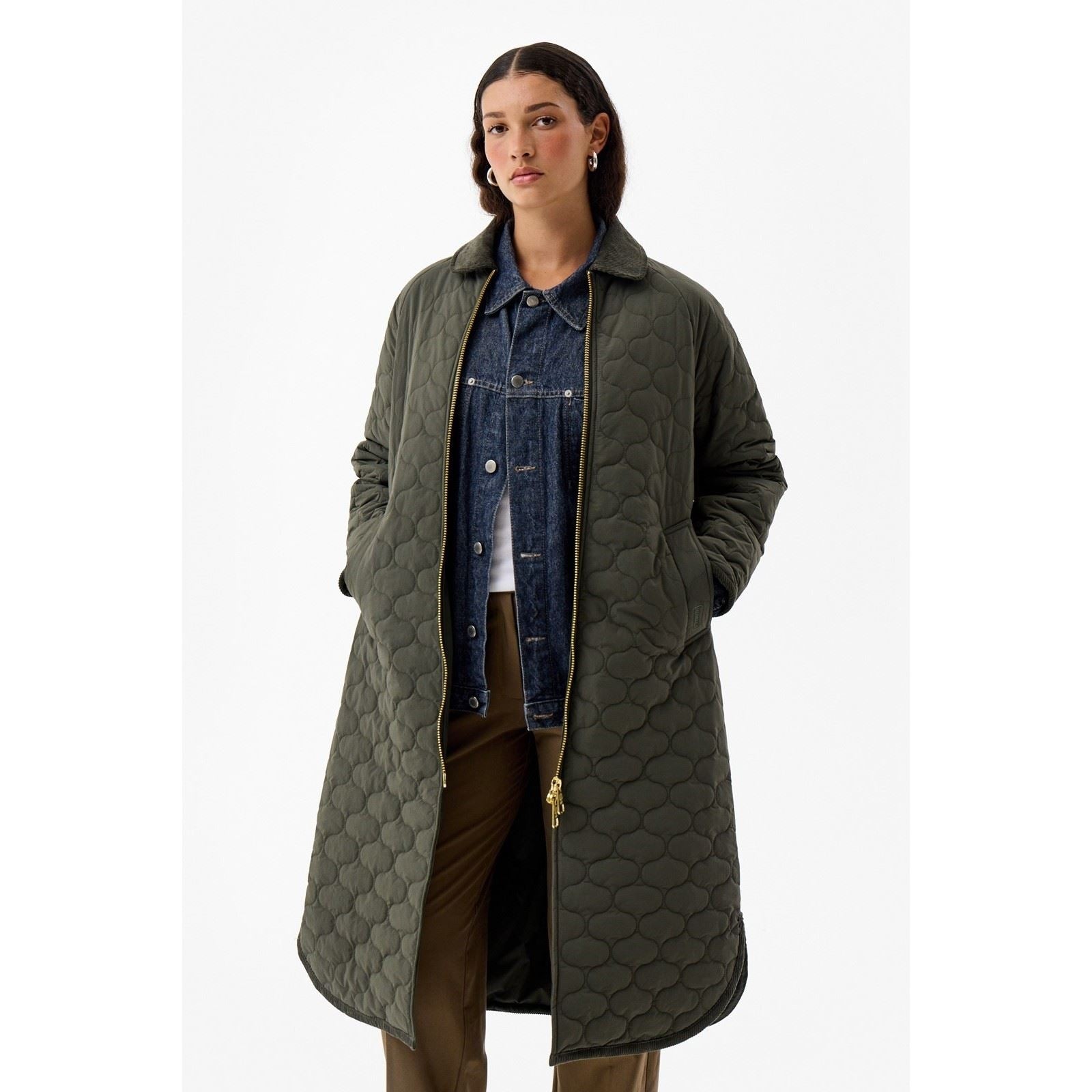 Long green quilted jacket by Hunter, featuring a zip front, side pockets, and a relaxed fit, layered over a denim jacket and styled with neutral pants.