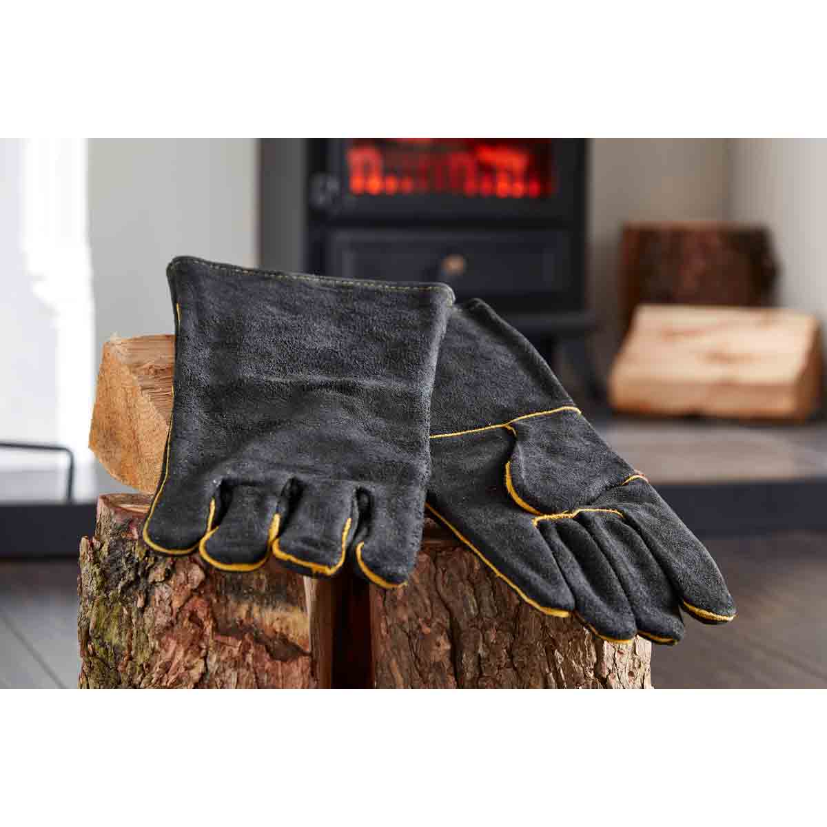 Black heat-resistant gloves with yellow stitching, made from durable material, displayed on a wooden log near a cozy fireplace setting.