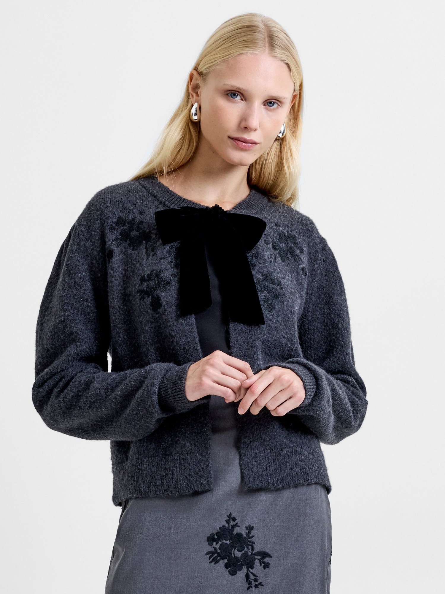Gray velvet cardigan featuring a front bow, floral embroidery, and a relaxed fit, styled with a model wearing a matching gray skirt.