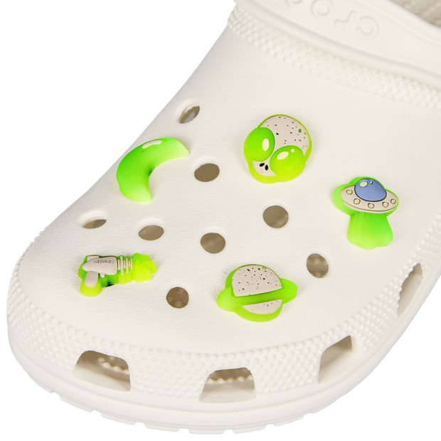 White Crocs featuring five glow-in-the-dark space-themed charms: a green alien, a crescent moon, a UFO, a planet, and a rocket, all set against the shoe's perforated design.