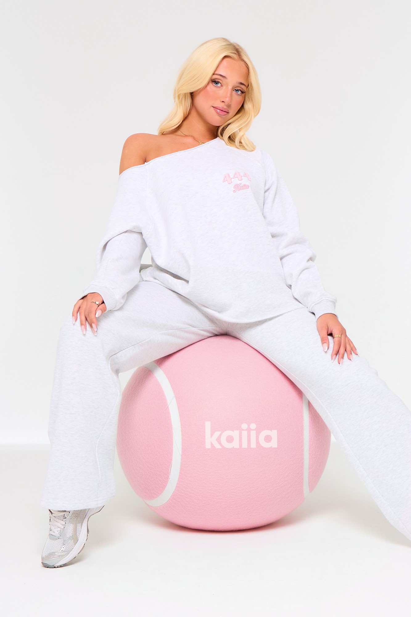 Light grey marl off-the-shoulder sweatshirt with pink "Kaiia" logo, paired with matching grey pants, model sitting on a pink oversized ball.