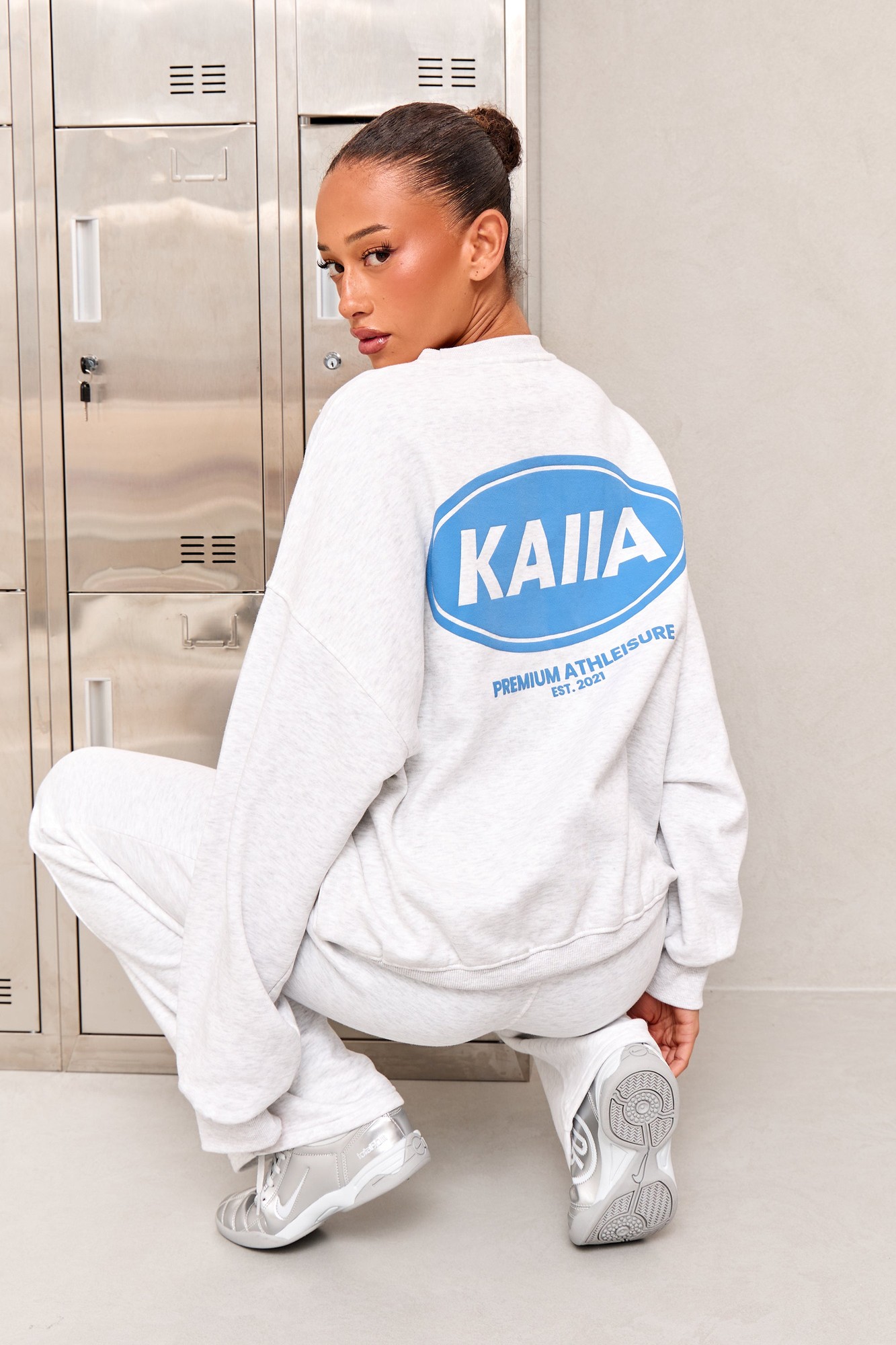 Light grey marl oversized sweatshirt featuring a vibrant blue "Kaiia" logo on the back, styled with matching sweatpants and silver sneakers against a minimalist backdrop.
