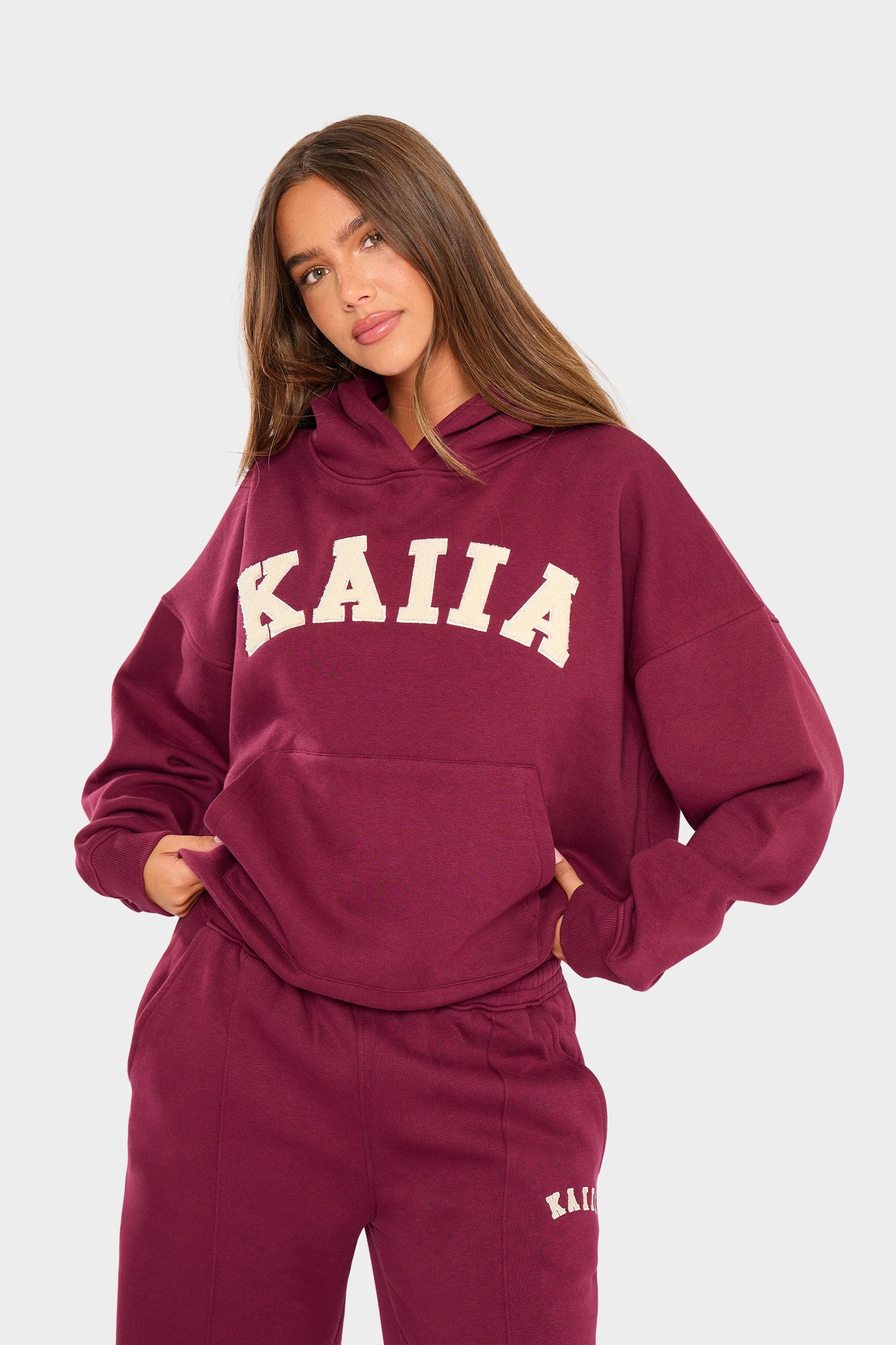 Burgundy oversized hoodie featuring "KAIIA" in cream letters, with a front pocket and a relaxed fit, styled with matching burgundy pants.