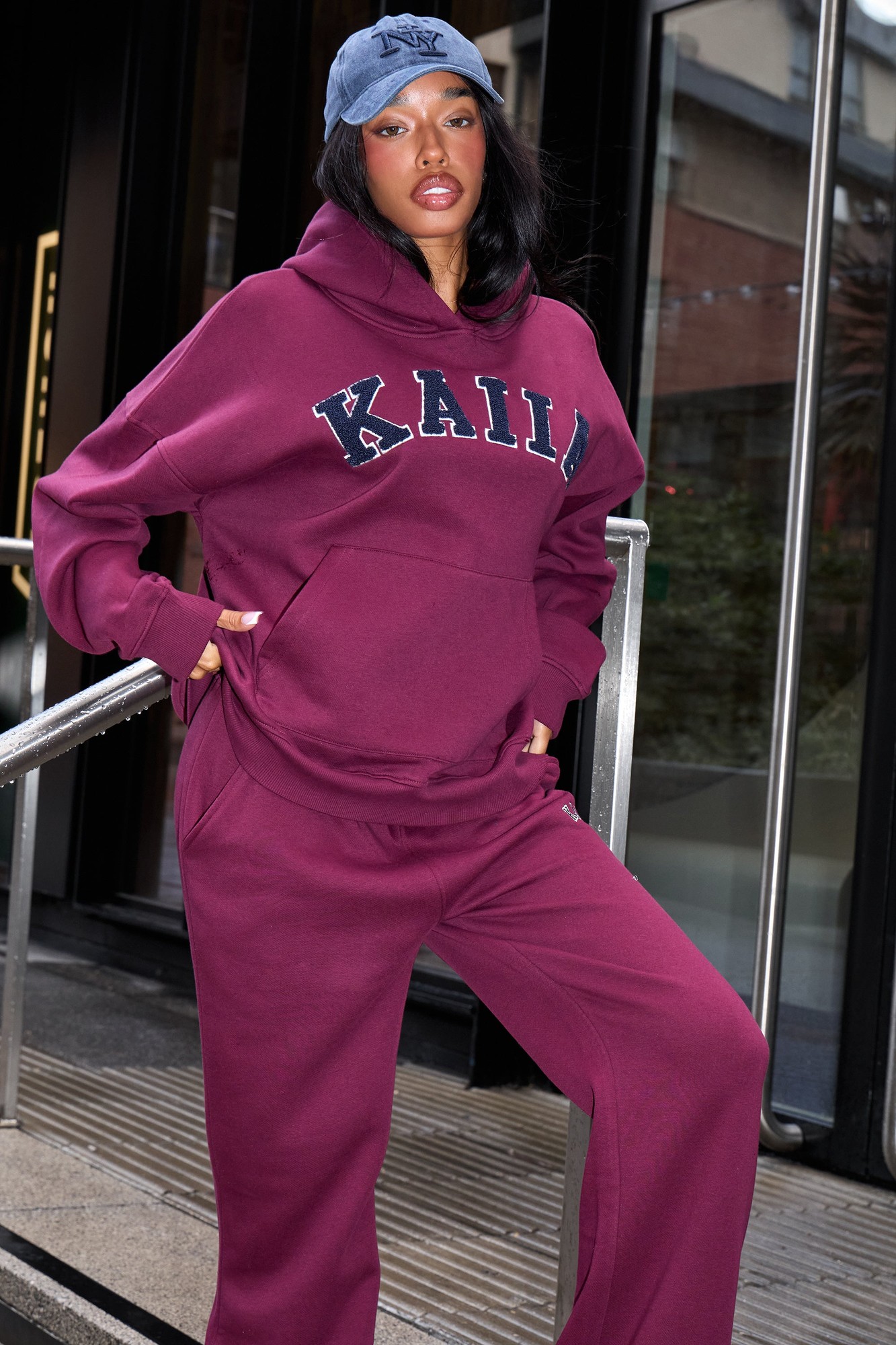 Burgundy oversized hoodie featuring "KAIIA" in navy letters, paired with matching pants, worn by a model with a blue cap, standing on stairs outside.